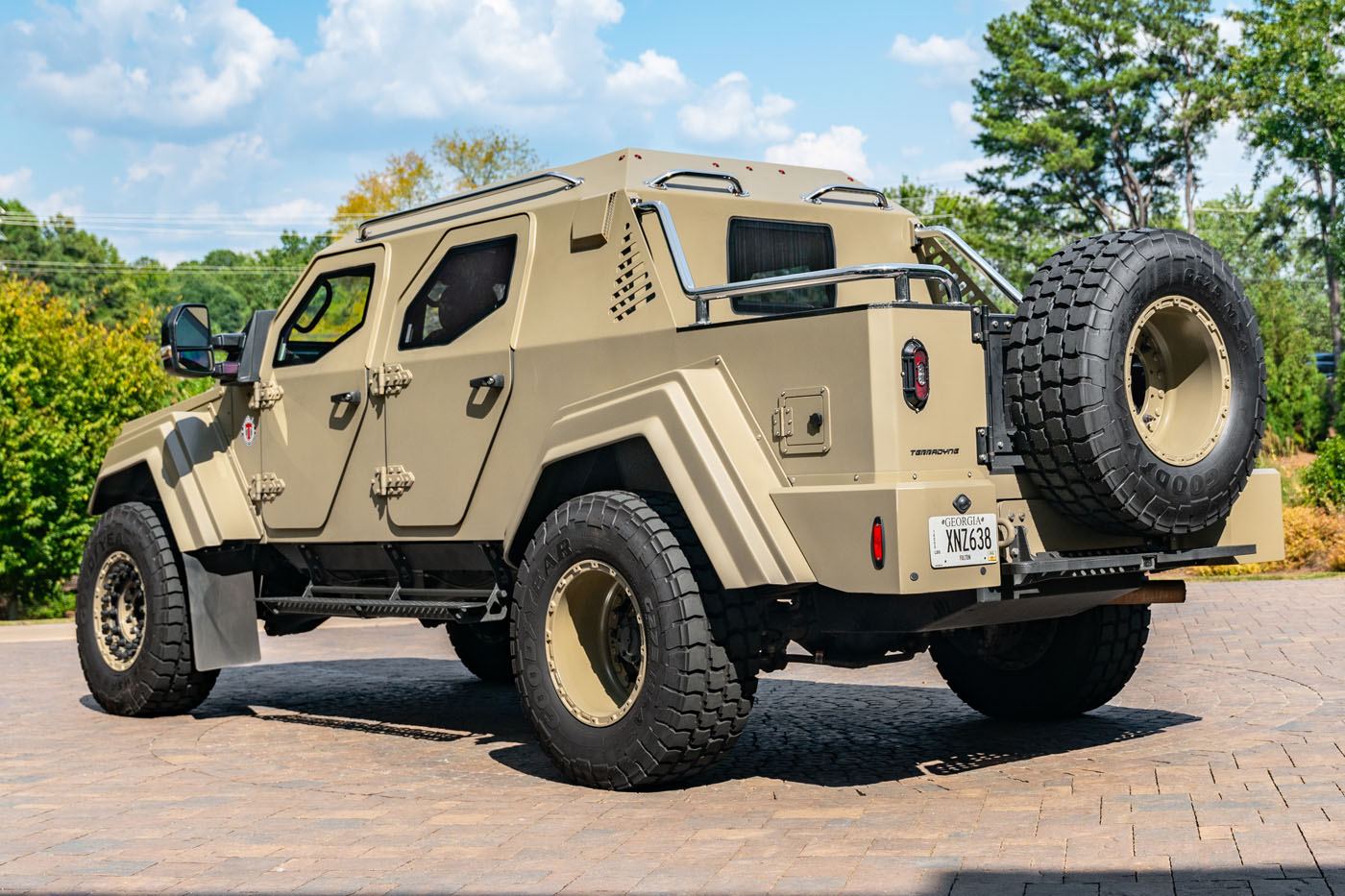 A tan Terradyne Gurkha armored vehicle with rugged tires and a rear-mounted spare tire is parked on a paved driveway, surrounded by greenery and trees, showcasing its street-legal versatility.