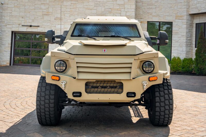 A beige Terradyne Gurkha fortress, fully street-legal, is parked on a brick driveway in front of a modern stone building.