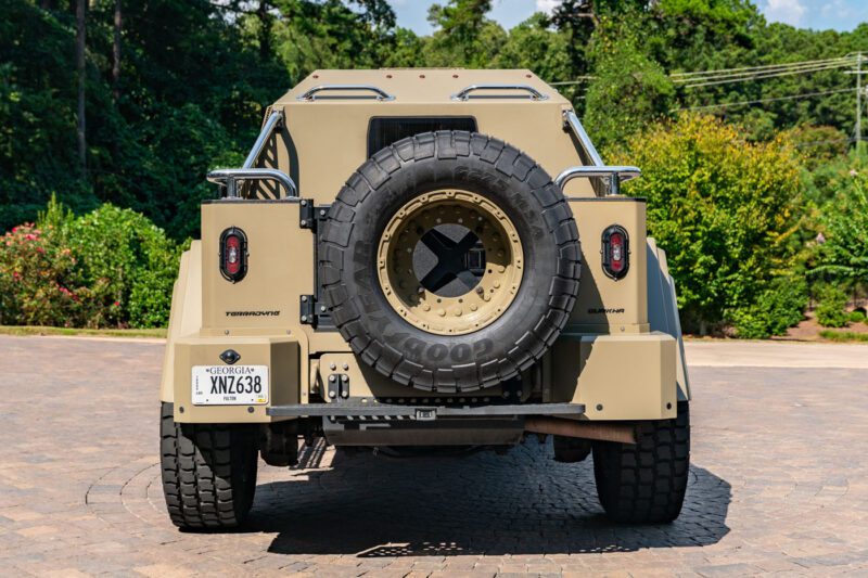 Rear view of a tan, rugged Terradyne Gurkha off-road vehicle with a spare tire mounted on the back, parked on a stone driveway with greenery in the background. This street-legal powerhouse is ready for any adventure.