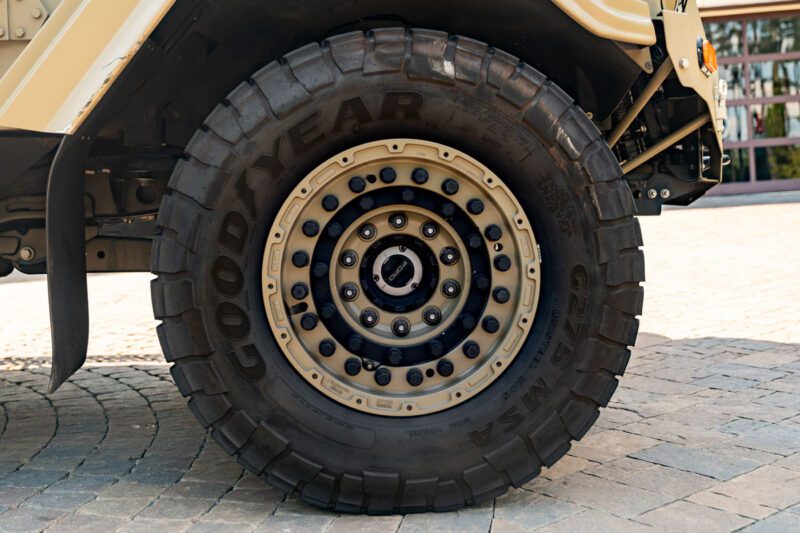 Close-up view of a large Goodyear off-road tire mounted on a beige Terradyne Gurkha, showing detailed treads and wheel bolts on a cobblestone surface. This impressive vehicle is street-legal and ready for sale.
