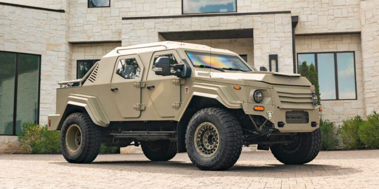 A tan 2020 Gurkha, a street-legal armored vehicle, is parked on a stone driveway in front of a modern light-colored building with large windows.
