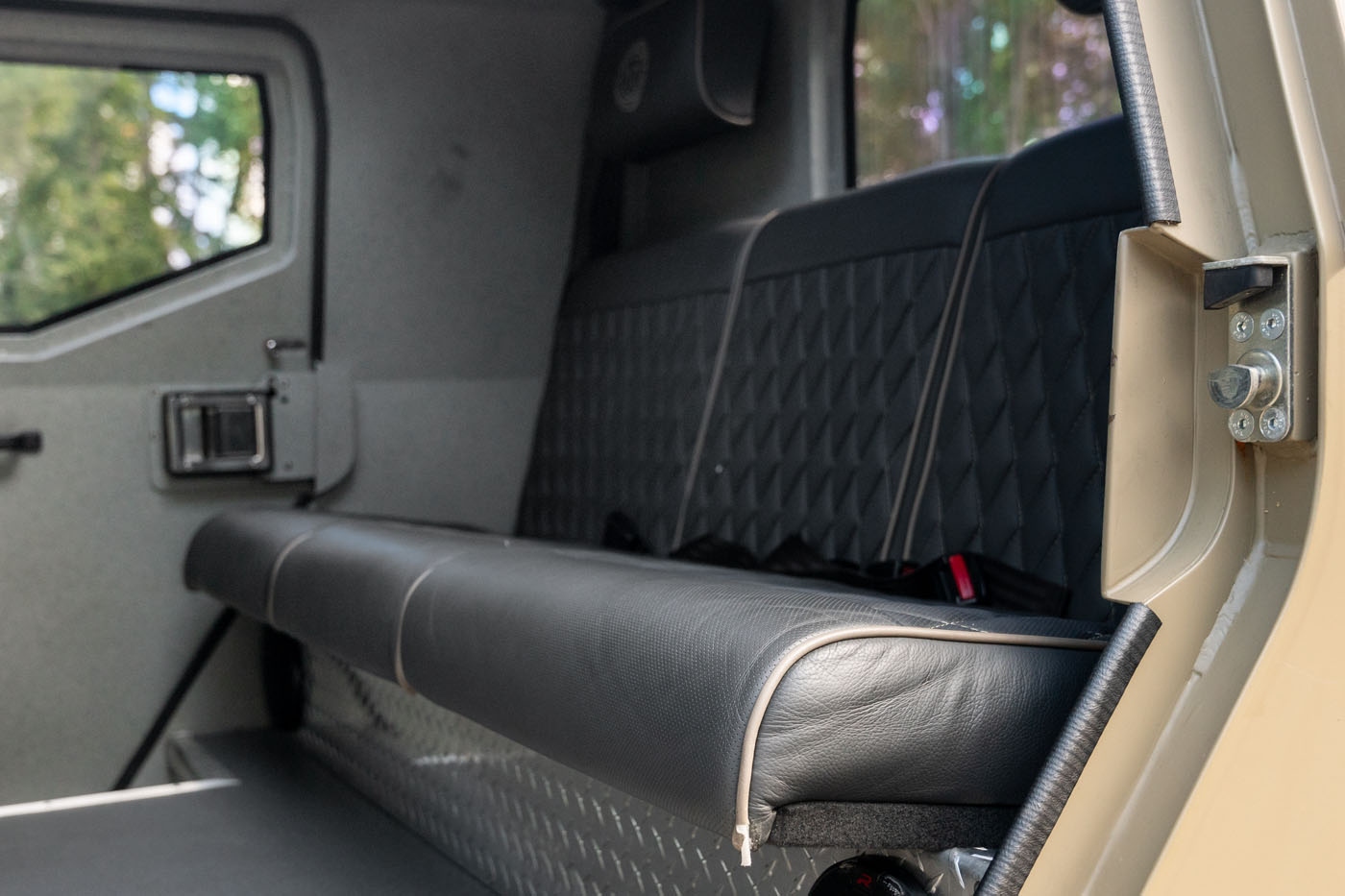 Black leather bench seat with diamond-pattern stitching inside a street-legal armored vehicle, viewed from the open side door, with a window and door latch visible in the background.