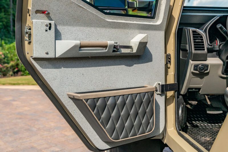 Close-up of a Terradyne Gurkha’s interior driver-side door panel featuring a textured surface, quilted black pocket, and visible handle, with part of the dashboard and steering wheel in view—this street-legal vehicle is now for sale.