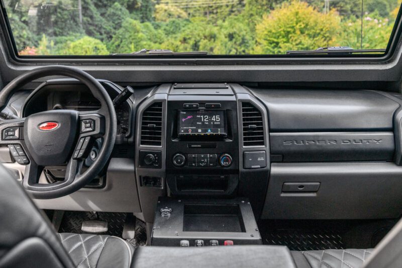 Interior view of a street-legal Terradyne Gurkha Super Duty truck cab showcasing the steering wheel, dashboard, central console with digital clock, and sleek black leather seats.
