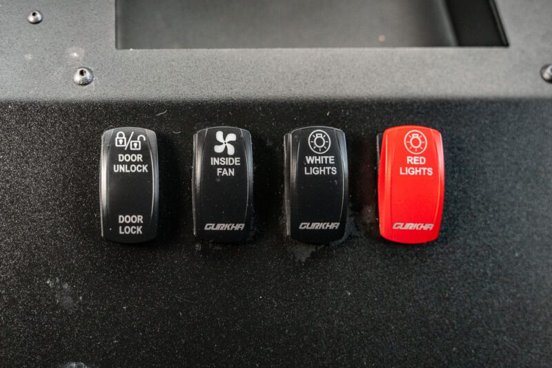 Four labeled toggle switches on this Terradyne Gurkha for sale panel: door unlock/lock, inside fan, white lights, and a distinctive red lights fortress switch; the red one stands out while the others are street-legal black.