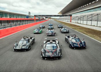 A group of high-performance sports cars is lined up on a race track with grandstands and empty seating visible in the background.