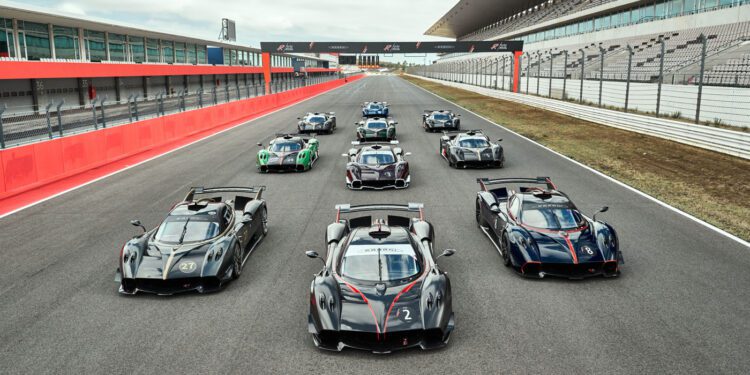 A group of high-performance sports cars is lined up on a race track with grandstands and empty seating visible in the background.