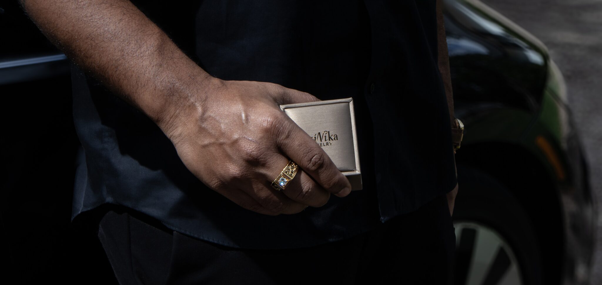 A person wearing a gold ring holds a small beige box labeled “Mika Jewelry,” showcasing rare legacies, while standing next to a black car.