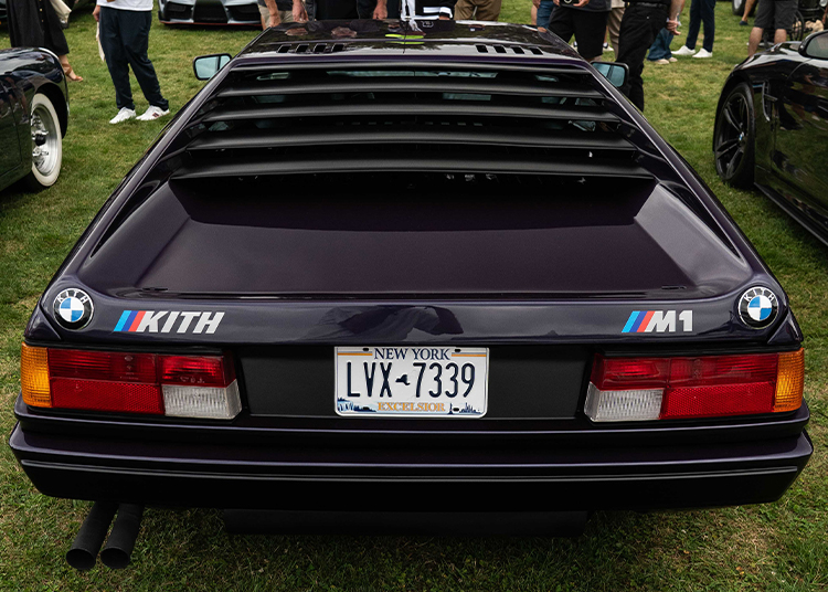 Rear view of a dark BMW M1 showcasing automotive art and exclusivity, with New York license plate, Kith and BMW logos, parked on grass at Bridge IX amid people and other cars.