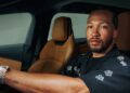 A man with a beard sits in the driver’s seat of a car with tan leather interior, wearing a black shirt and a wristwatch, reminiscent of Josh Hart in a sleek Lucid campaign.