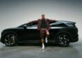 A man wearing sunglasses and a matching maroon outfit leans against a sleek black SUV in a studio setting, channeling Josh Hart’s effortless style in this Lucid campaign.