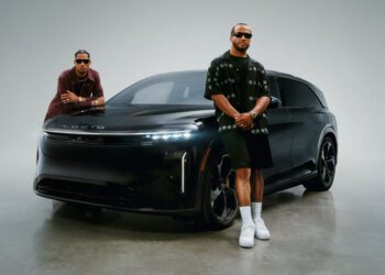 Jalen Brunson and Josh Hart, both wearing sunglasses, stand in front of a black Lucid electric vehicle in a studio setting for a Lucid campaign.
