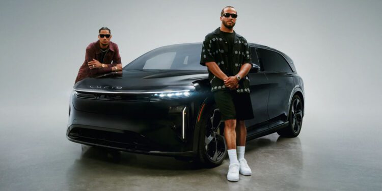 Jalen Brunson and Josh Hart, both wearing sunglasses, stand in front of a black Lucid electric vehicle in a studio setting for a Lucid campaign.