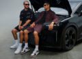 Two men wearing sunglasses and casual outfits, reminiscent of Jalen Brunson’s laid-back style, sit on the front of a black car with its hood open in a plain studio setting.