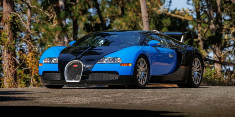 A blue and black Bugatti Veyron sports car is parked on a paved road with trees in the background, ready to turn heads at Broad Arrow’s Las Vegas 2025 event.