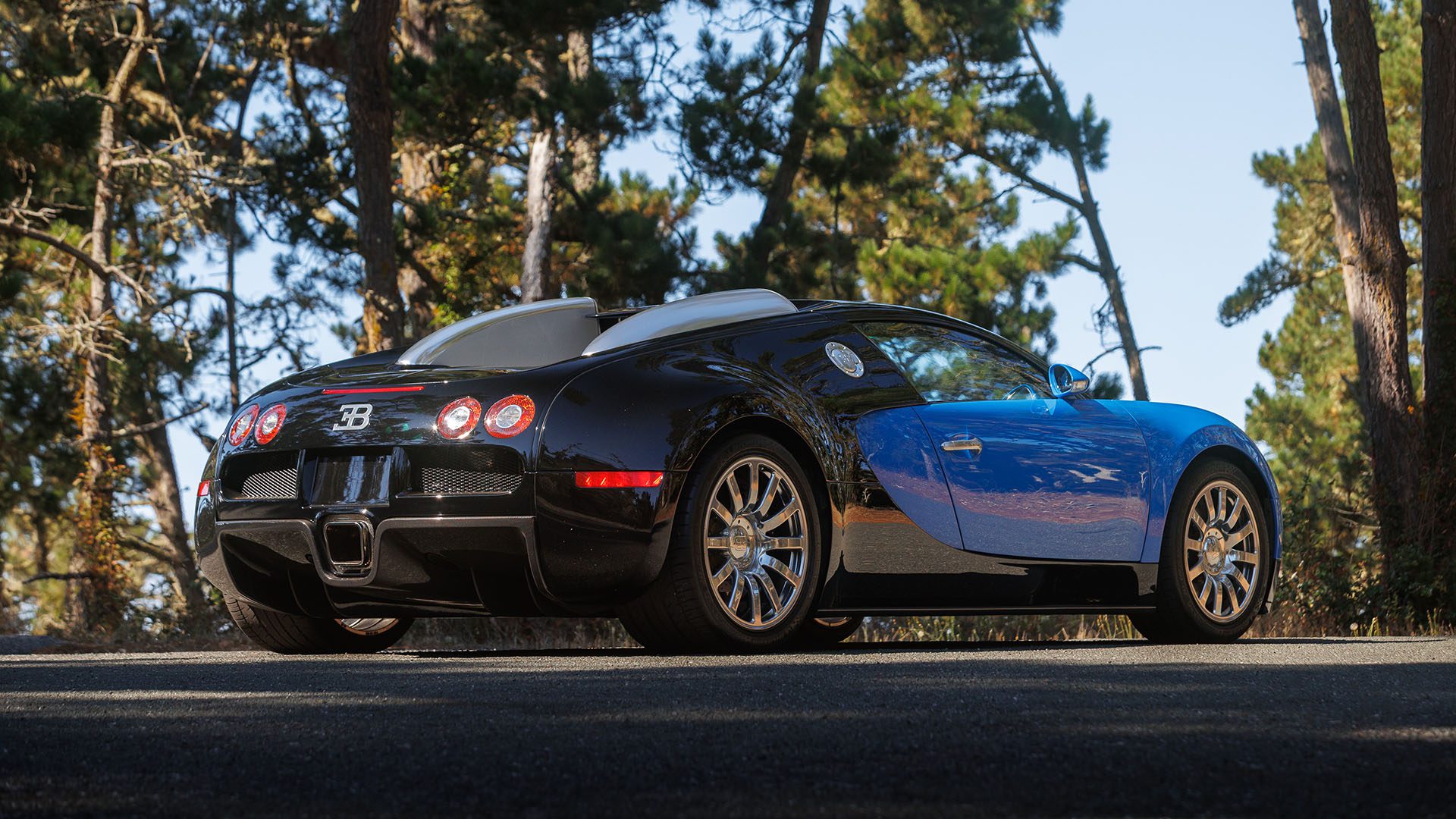 A black and blue Bugatti Veyron, set to impress at Broad Arrow’s Las Vegas 2025 event, is parked on a paved road with trees in the background.