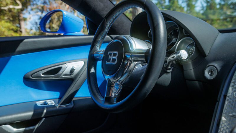 Interior view of a luxury car, showcasing the sleek Bugatti-branded steering wheel, dashboard, and blue door panel of a 2010 Bugatti Veyron, featured at Broad Arrow Las Vegas 2025.