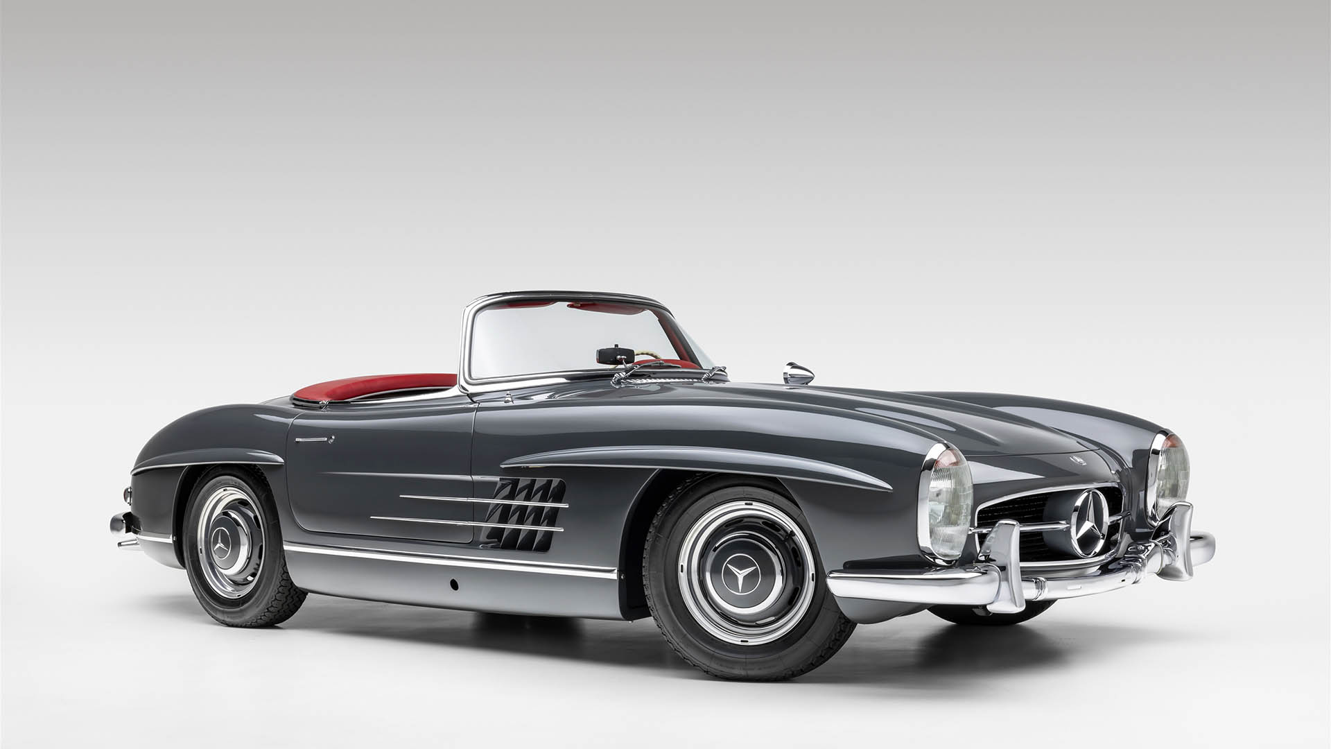 A classic dark grey 2009 Mercedes-Benz SLR McLaren convertible with a red interior is displayed in a studio setting with a white background, set to appear at Broad Arrow Las Vegas 2025.