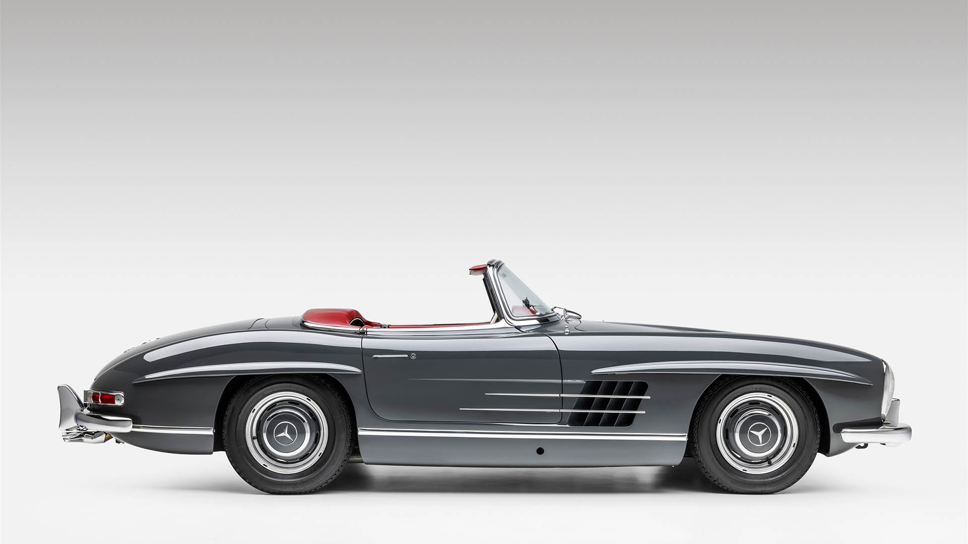 A classic gray Mercedes-Benz 300SL convertible with a red interior, viewed from the side against a plain white background, evokes the timeless elegance showcased at events like Broad Arrow Las Vegas 2025.