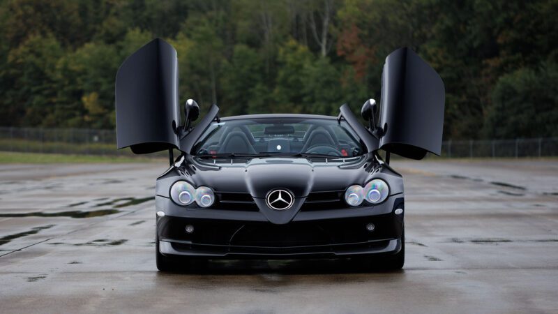 A black Mercedes-Benz SLR McLaren with its gullwing doors open sits on wet pavement, surrounded by trees—a striking sight reminiscent of Broad Arrow's Las Vegas 2025 showcase.