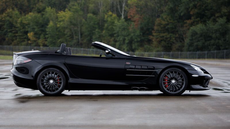 A black Mercedes-Benz SLR McLaren convertible with its top down is parked on wet pavement, trees and a fence in the background—a striking sight reminiscent of Broad Arrow’s upcoming Las Vegas 2025 showcase.