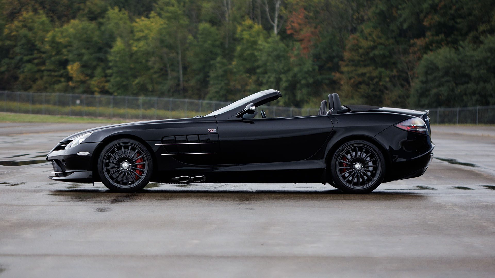 A 2009 Mercedes-Benz SLR McLaren convertible sports car is parked on wet pavement with trees and a fence in the background, ready to impress at Broad Arrow Las Vegas 2025.