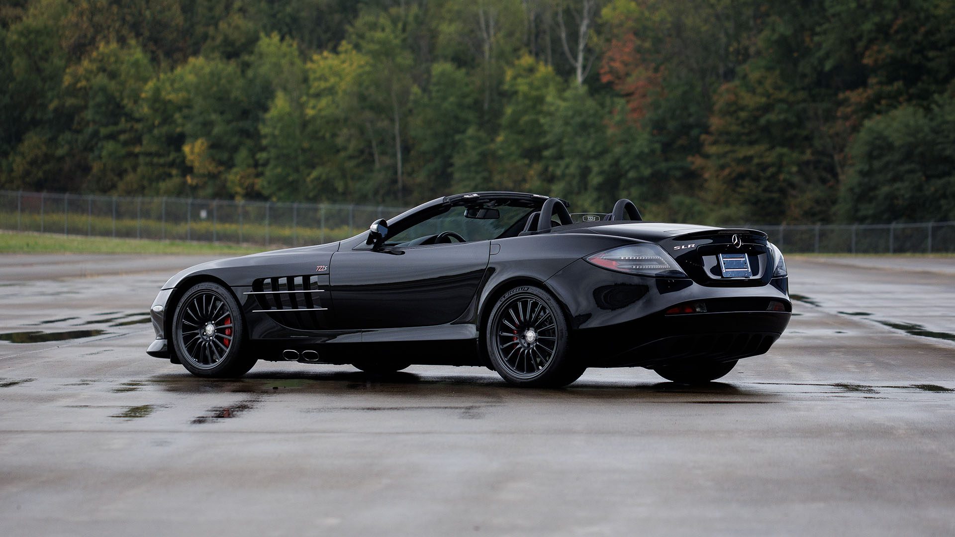 A sleek black Mercedes-Benz SLR McLaren convertible sits parked on a wet surface, framed by trees and a fence—an eye-catcher for Broad Arrow’s Las Vegas 2025 event.