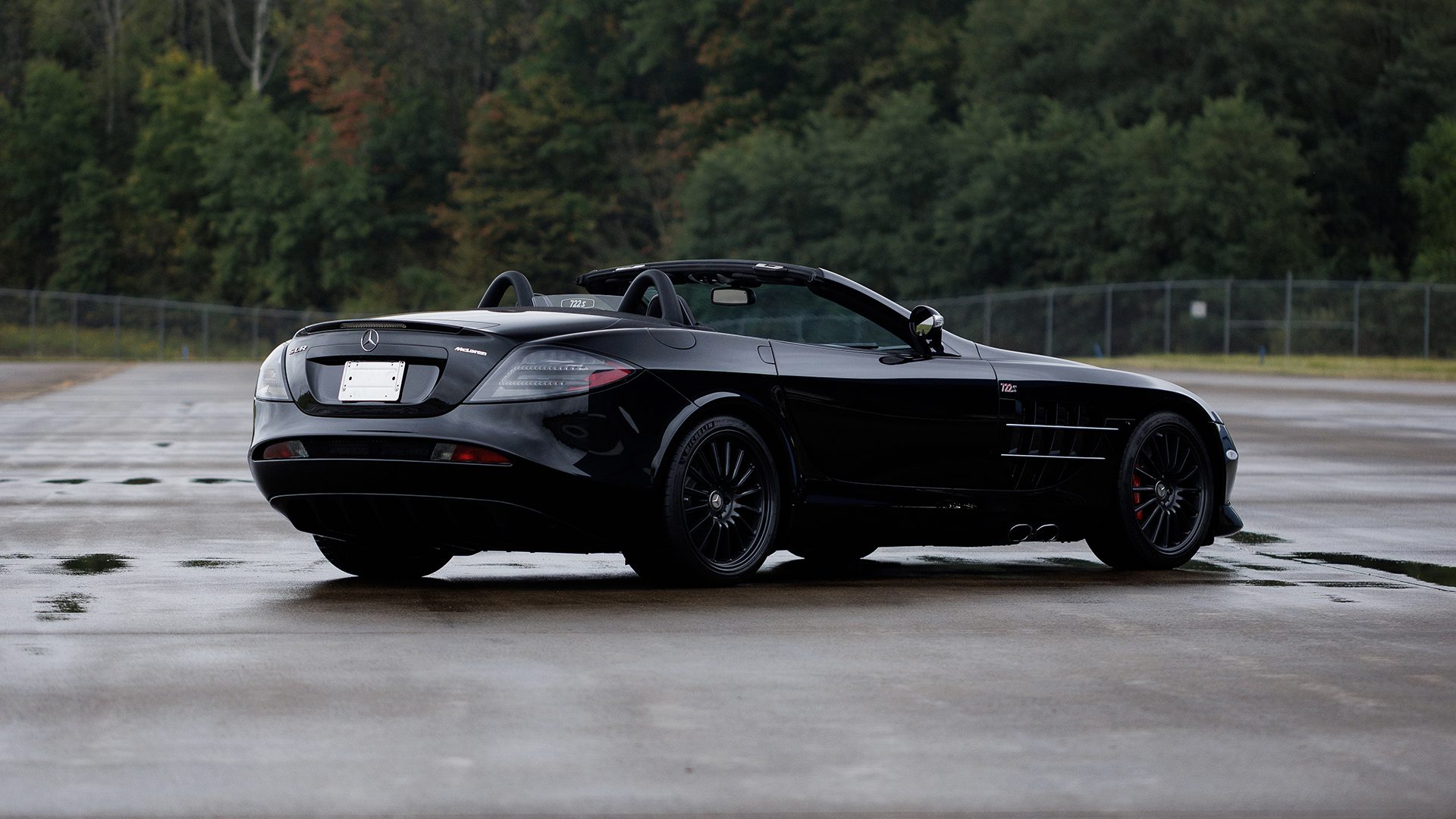 A black Mercedes-Benz convertible is parked on wet pavement with a wooded area and chain-link fence in the background, reminiscent of sleek vehicles showcased at Broad Arrow's Las Vegas 2025 event.