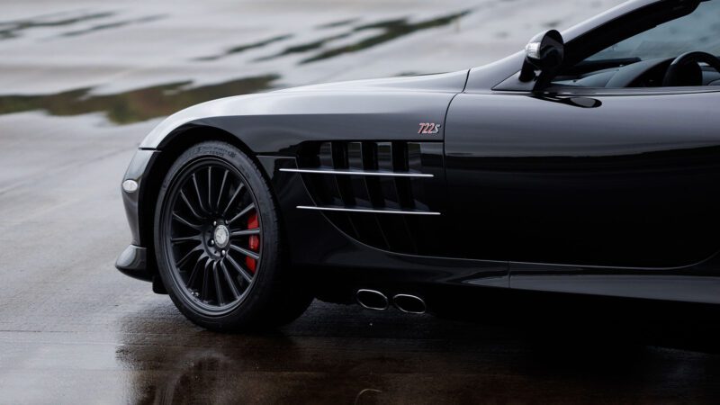 Close-up of the front left side of a black sports car with "722 S" badge, black wheels, and red brake caliper, parked on a wet surface at Broad Arrow’s Las Vegas 2025 event.
