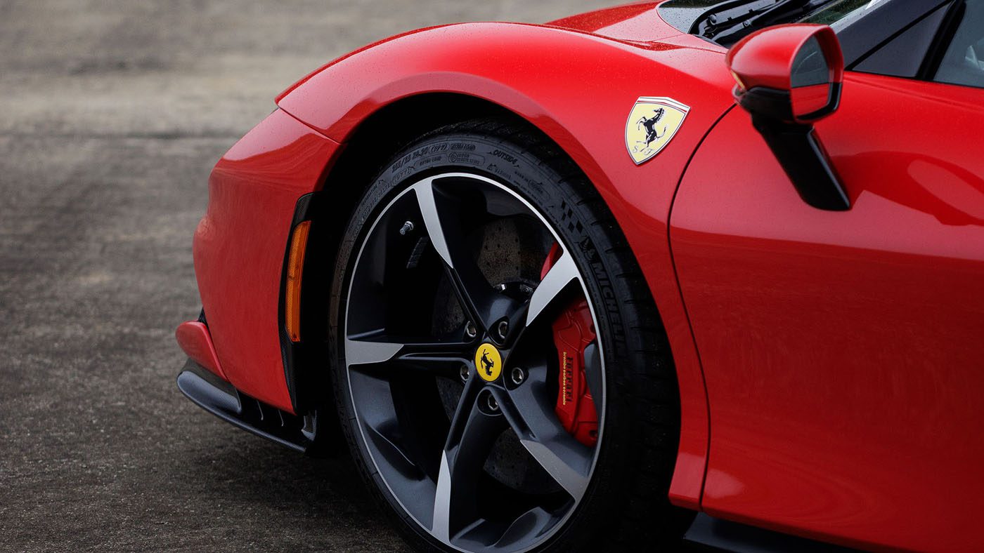 Close-up of the front left wheel and fender of a red Ferrari sports car, showing the Ferrari logo and black alloy wheel with red brake caliper—captured at Broad Arrow Las Vegas 2025 alongside icons like the 2010 Bugatti Veyron.