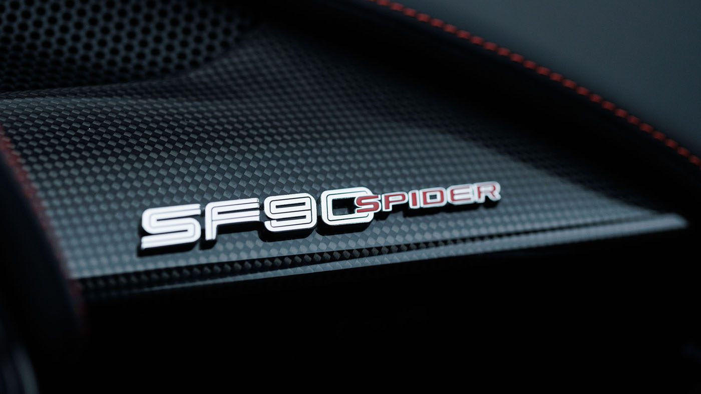 Close-up of a car interior panel featuring the "SF90 Spider" badge in silver and red lettering on a textured carbon fiber surface, reminiscent of the luxurious detailing found in a 2010 Bugatti Veyron.