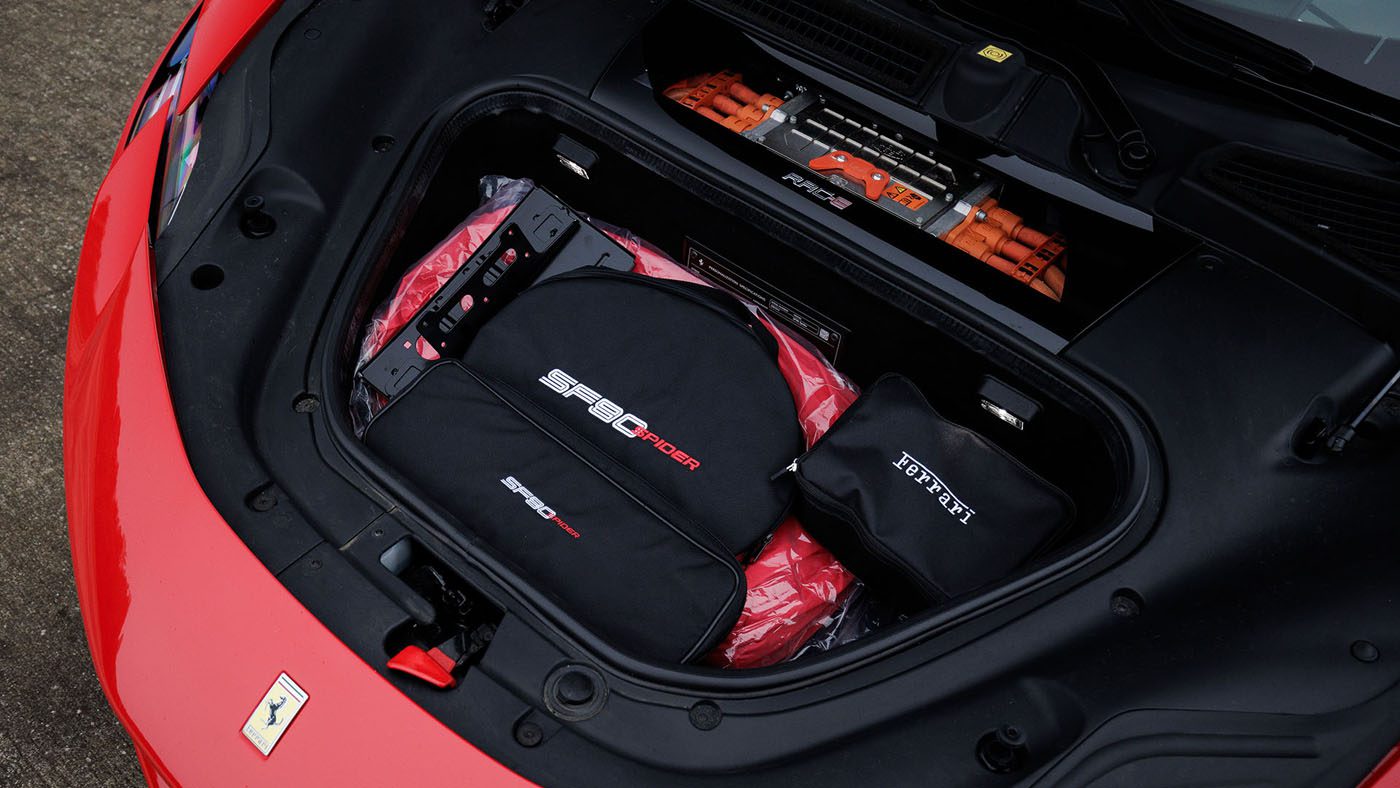 The front trunk of a Ferrari SF90 features branded luggage, a carrying case, and visible orange high-voltage components—just one highlight at the Broad Arrow Las Vegas 2025 Preview, where icons like the 2010 Bugatti Veyron will also be showcased.