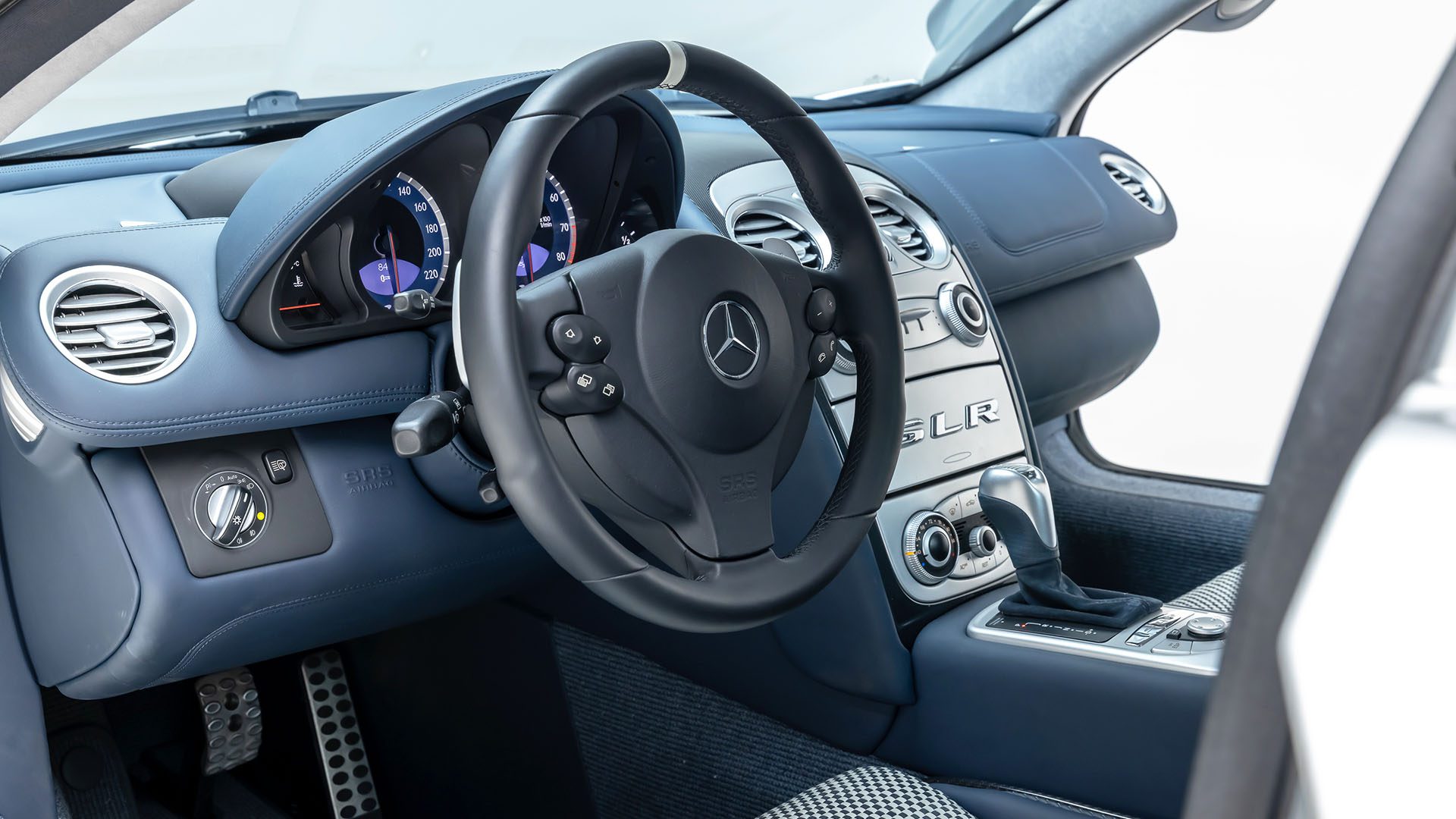 Close-up view of a Mercedes-Benz SLR McLaren interior showing a black steering wheel, instrument cluster, center console, and blue leather dashboard at Broad Arrow Las Vegas 2025.