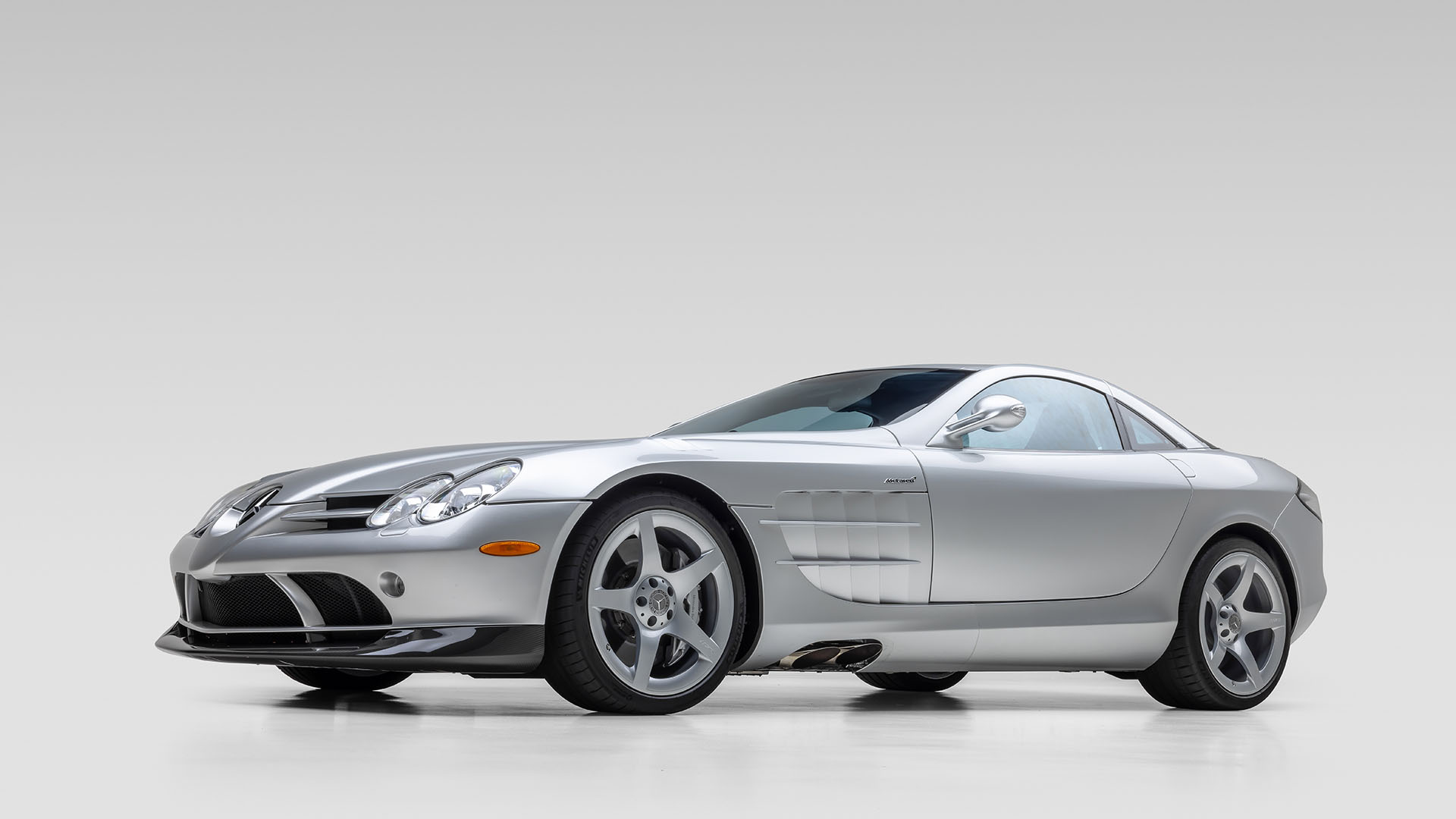 A silver Mercedes-Benz SLR McLaren sports car, inspired by the legendary Mercedes-Benz 300 SL, is shown from a front side angle on a plain light gray background.