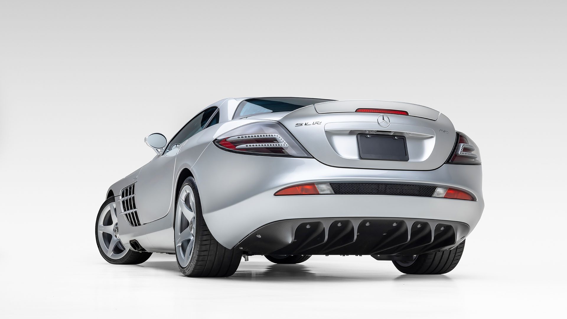 A silver Mercedes-Benz SLR McLaren sports car, reminiscent of the iconic Mercedes-Benz 300 SL Roadster, is viewed from the rear against a plain white background, hinting at the elegance of a Broad Arrow Las Vegas 2025 Preview.