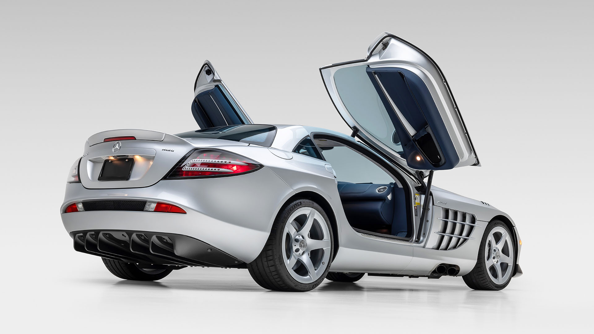 A silver Mercedes-Benz 300 SL Roadster with its gullwing doors open, viewed from the rear three-quarter angle against a plain white background—an icon worthy of the Broad Arrow Las Vegas 2025 Preview.