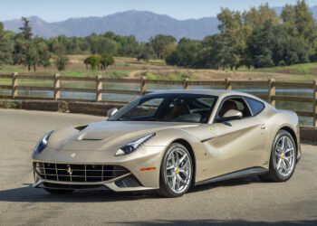 A beige Ferrari F12berlinetta 70th Anniversary Edition is parked on a paved road beside a wooden fence, with trees and mountains in the background, capturing a timeless scene worthy of Las Vegas 2025.