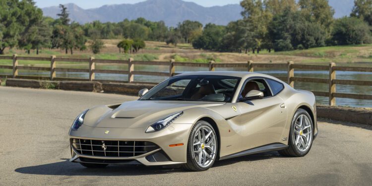 A beige Ferrari F12berlinetta 70th Anniversary Edition is parked on a paved road beside a wooden fence, with trees and mountains in the background, capturing a timeless scene worthy of Las Vegas 2025.