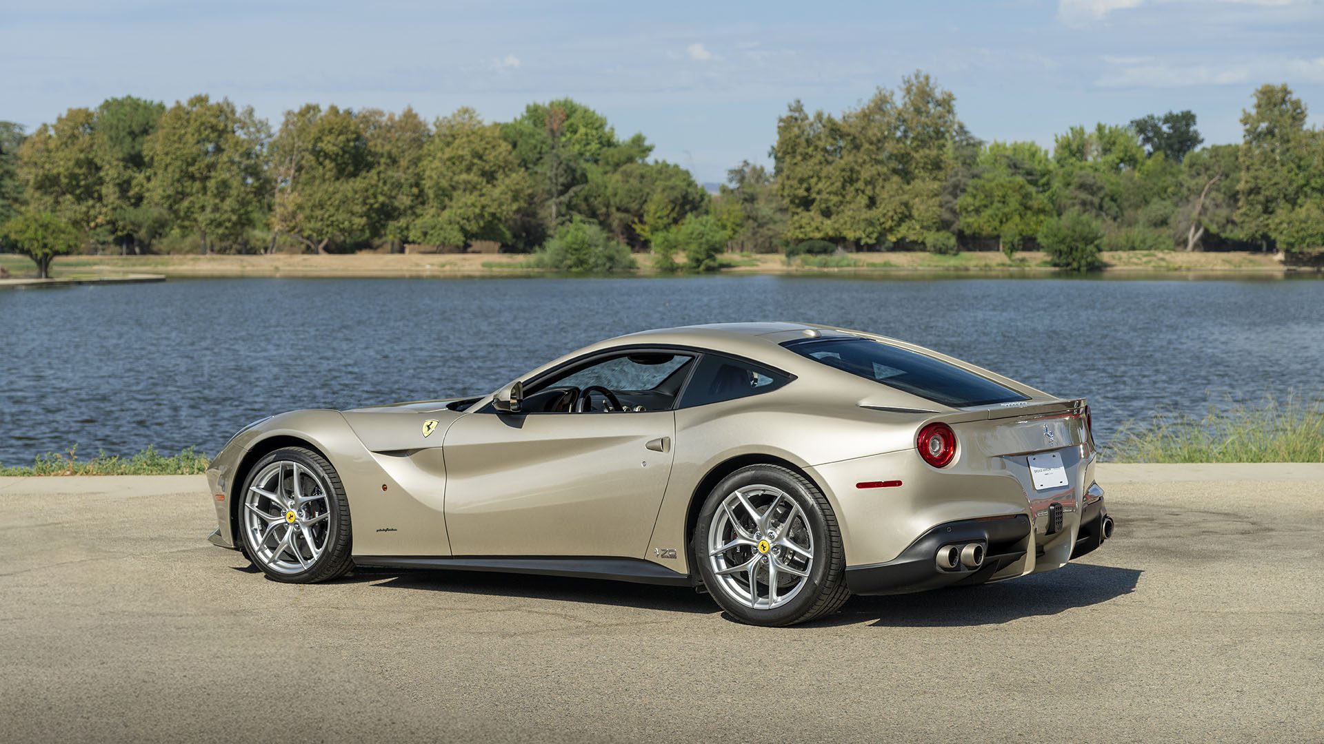 Metalinio smėlio spalvos Ferrari F12berlinetta kupė stovi asfaltuotoje vietoje prie ežero, o fone medžiai ir mėlynas dangus – stulbinantis 70-mečio leidimas, užfiksuotas prieš 2025 m. Broad Arrow Las Vegas.