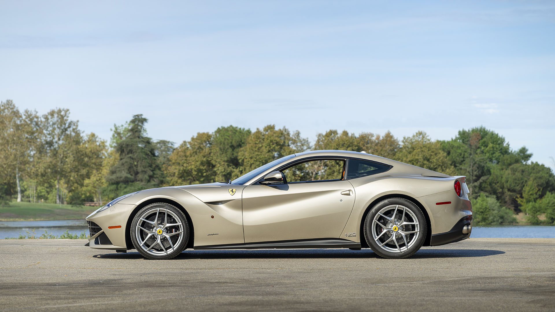 Sidabrinis sportinis automobilis Ferrari F12berlinetta stovi ant asfaltuotos dangos su medžiais ir vandens telkiniu fone po giedru dangumi, demonstruodamas savo eleganciją prieš 2025 m. Las Vegasą.