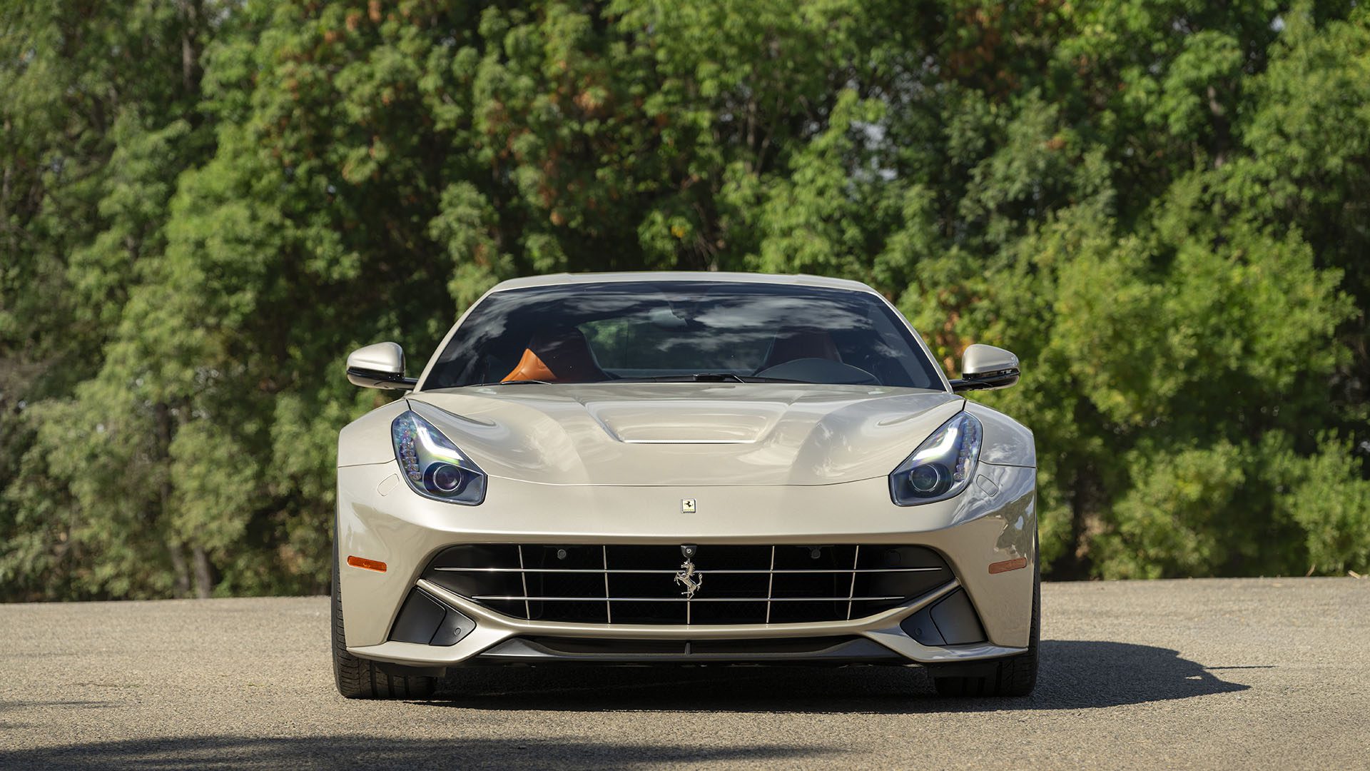 A front view of a beige Ferrari F12berlinetta 70th Anniversary Edition is parked on a paved road, surrounded by green trees, as seen at Broad Arrow Las Vegas 2025.