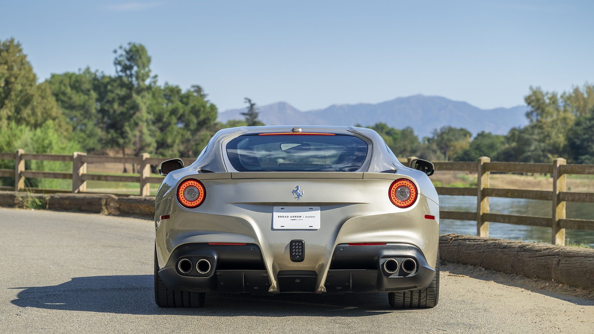 Sidabrinė „Ferrari F12berlinetta“ žiūrint iš galo, pastatyta ant kelio šalia medinės tvoros, su medžiais ir kalnais fone – ikoniška scena prieš „Broad Arrow Las Vegas 2025“.