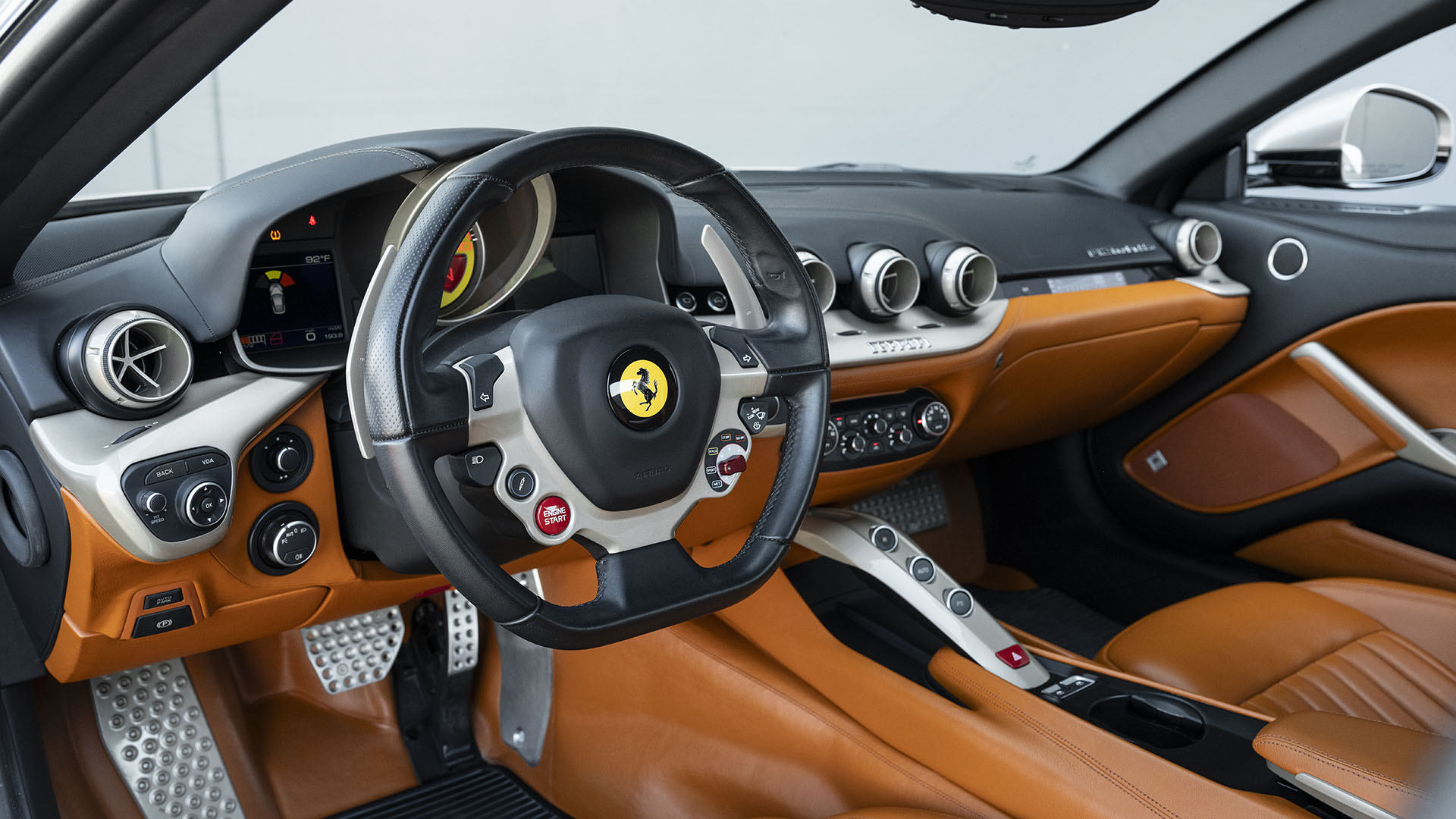 Prabangaus sportinio automobilio Ferrari F12berlinetta interjeras su rudos spalvos odinėmis sėdynėmis, juodu prietaisų skydeliu, 70-mečio leidimo ženkleliu ant vairo ir metaliniais akcentais ant valdiklių ir pedalų.