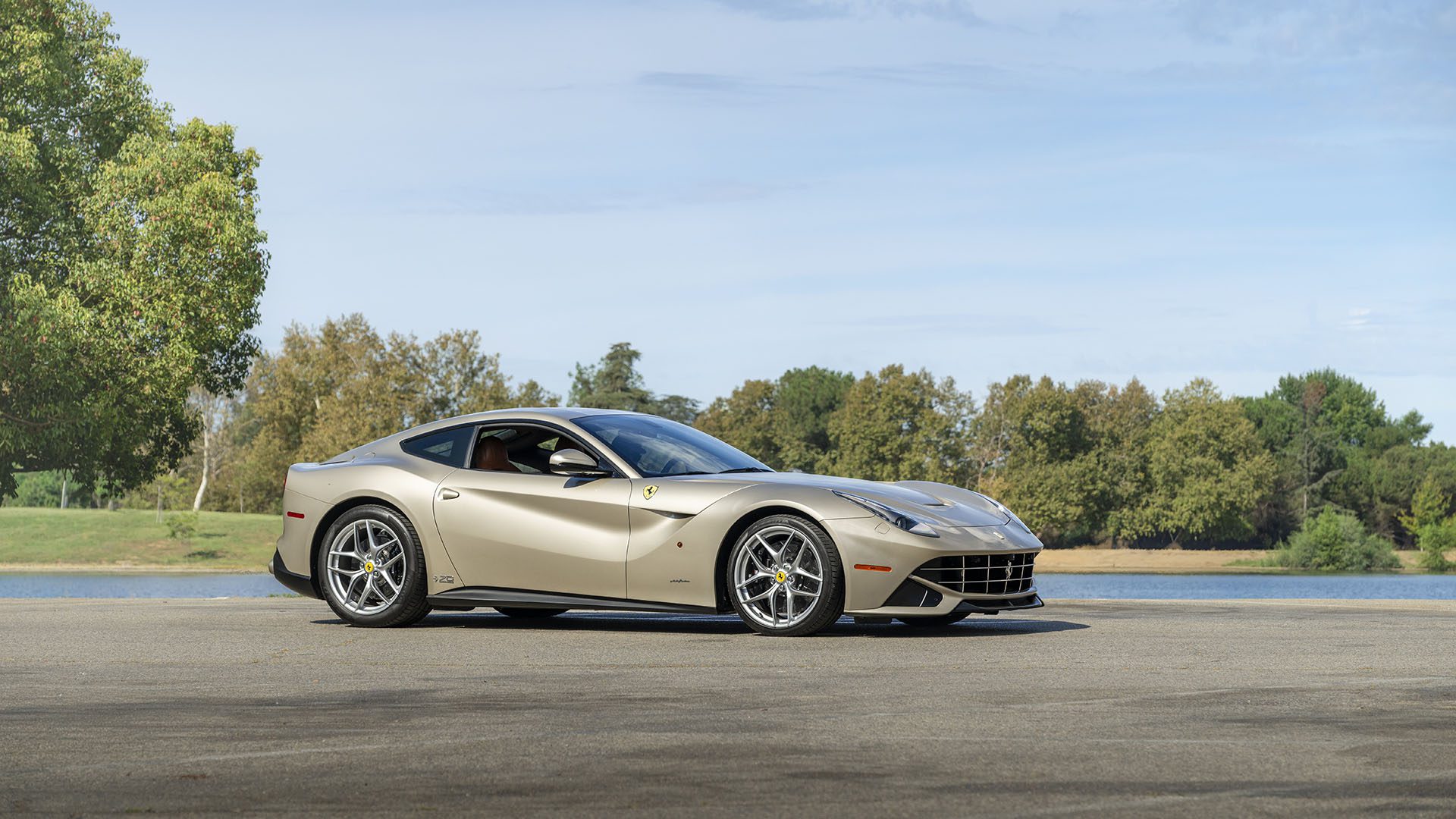 Sidabrinis „Ferrari F12berlinetta 70th Anniversary Edition“ pastatytas asfaltuotoje aikštelėje prie ežero, o fone medžiai ir mėlynas dangus – stulbinantis vaizdas prieš „Broad Arrow Las Vegas 2025“.
