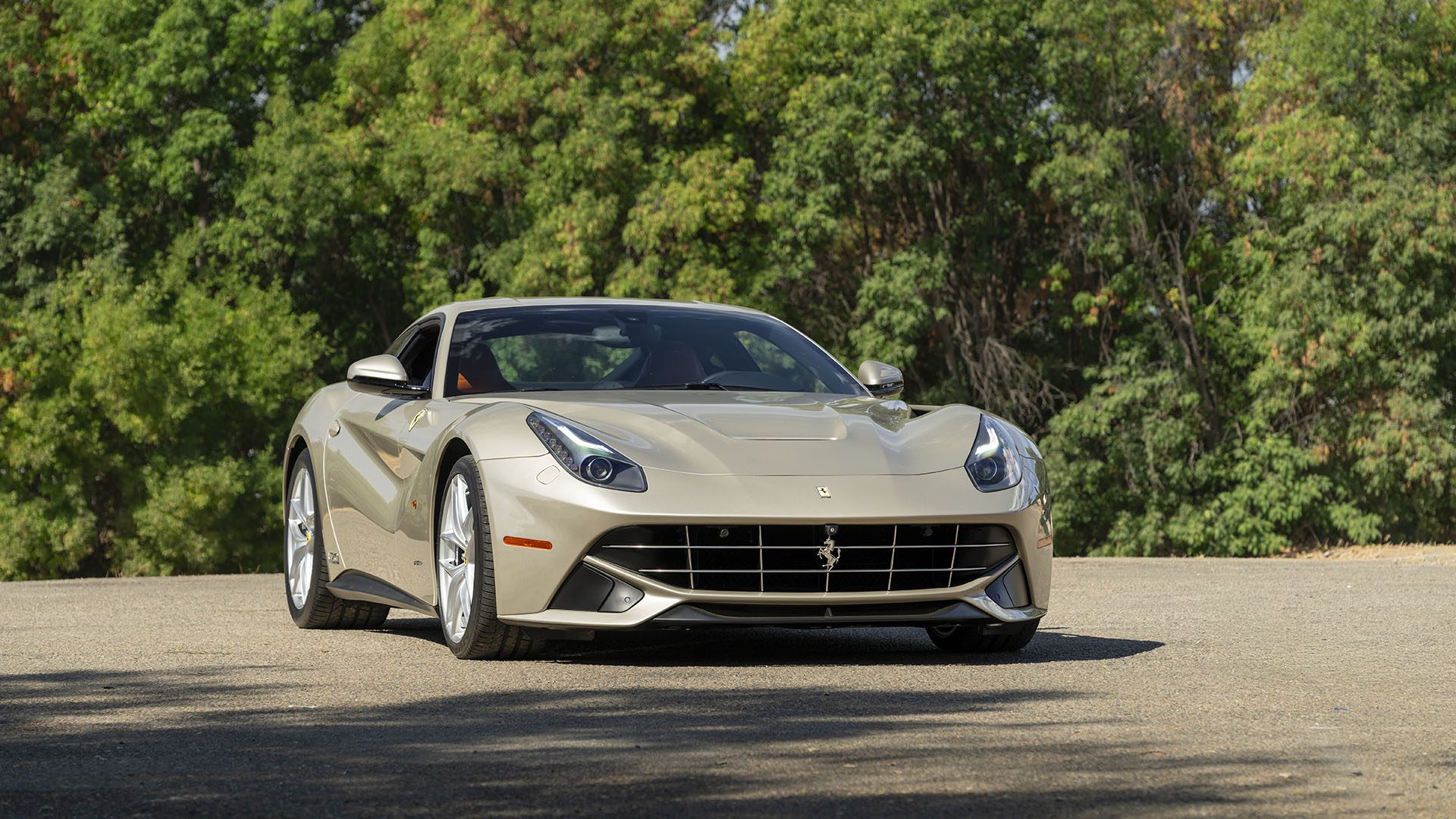 Sidabrinis Ferrari F12berlinetta 70th Anniversary Edition pastatytas ant asfaltuoto paviršiaus su žaliais medžiais fone.