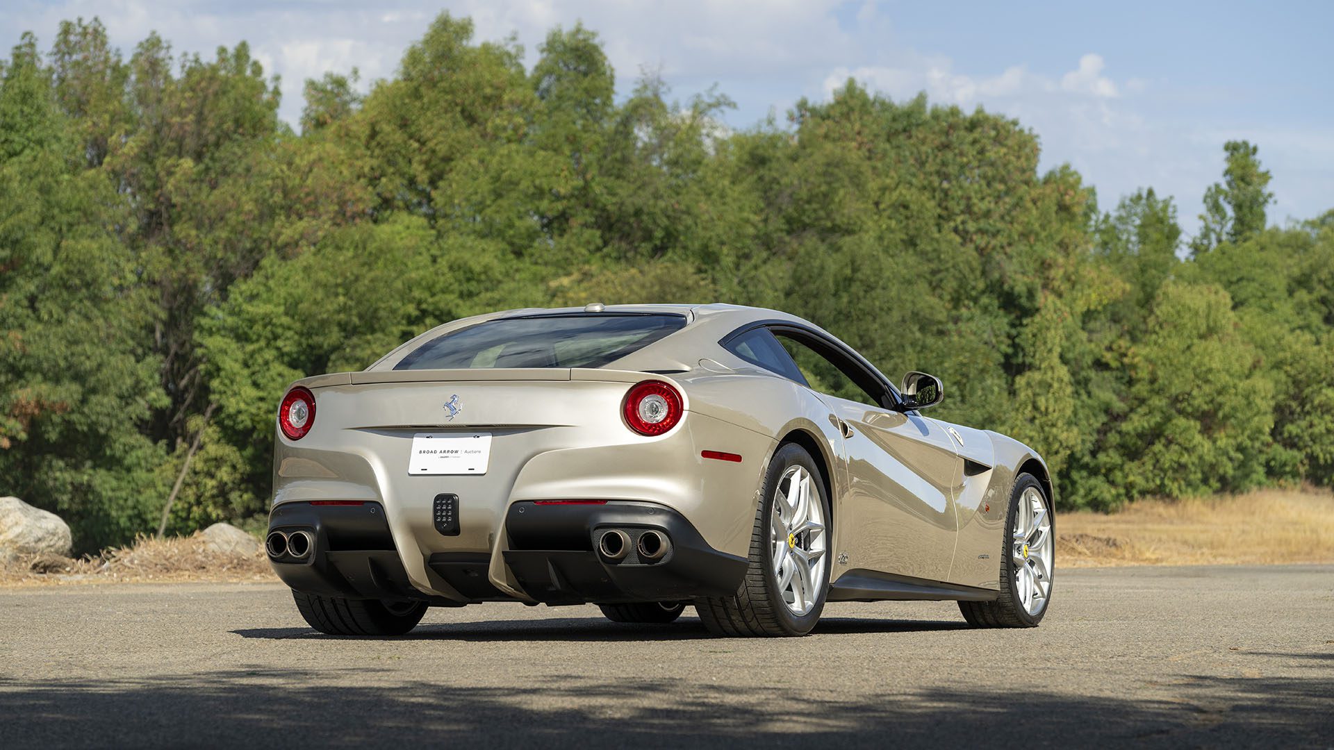 Sidabrinis „Ferrari F12berlinetta 70th Anniversary Edition“ pastatytas ant asfaltuoto kelio, apsuptas žalių medžių ir iš dalies debesuoto dangaus – tai puiki „Broad Arrow Las Vegas 2025“ scena.