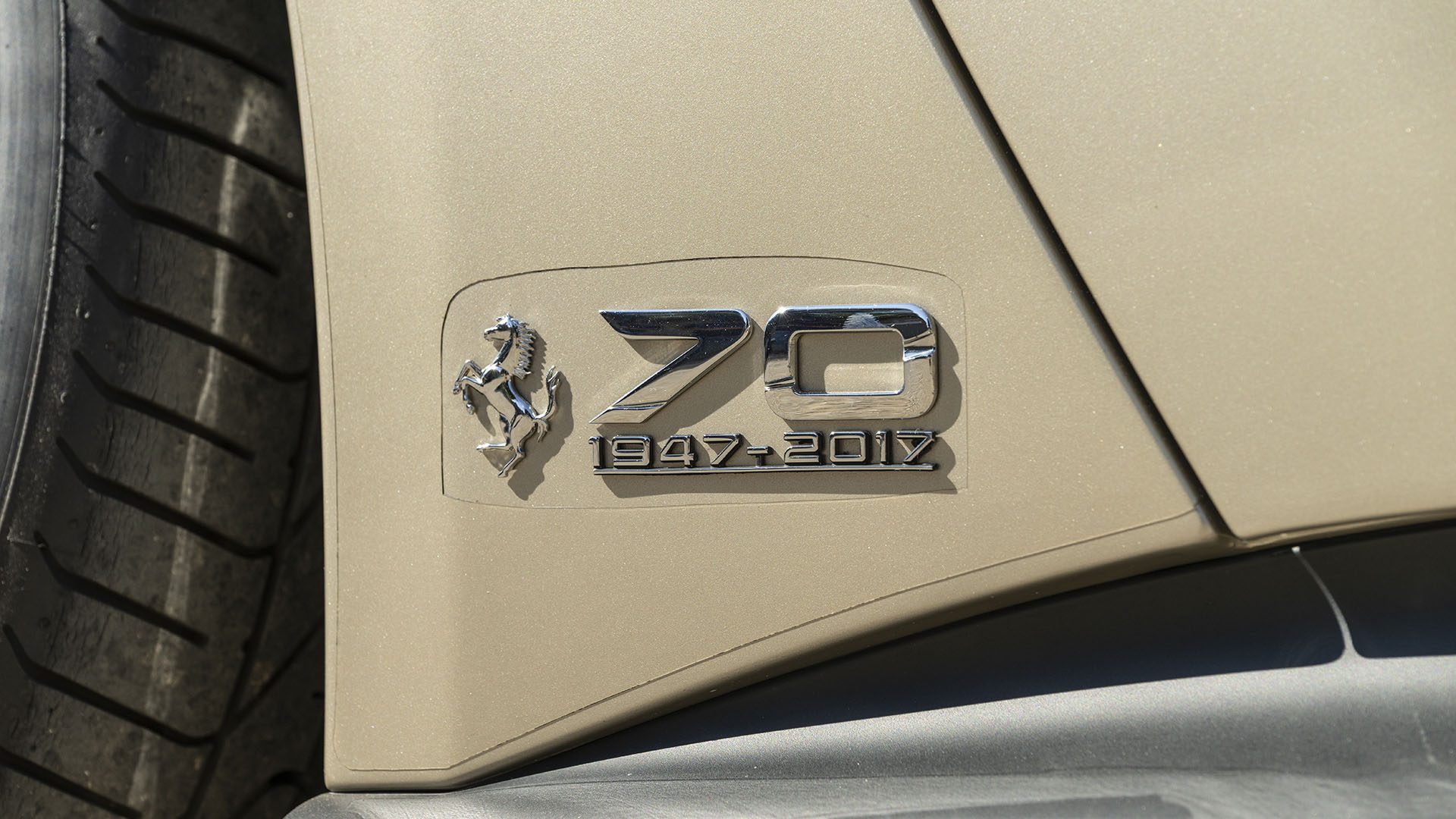 Ferrari F12berlinetta emblema iš arti "70" ir "1947-2017 m" smėlio spalvos automobilyje, švenčiančiame 70-ies metų jubiliejų – simbolinį akcentą prieš „Broad Arrow Las Vegas 2025“ renginį.