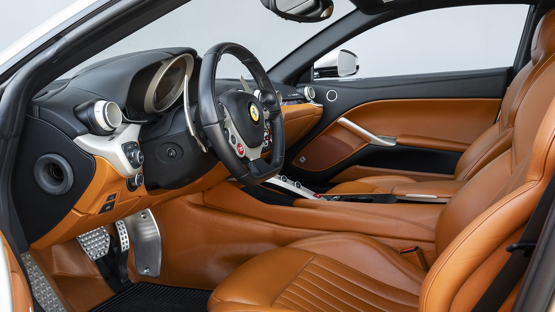 Vaizdas į „Ferrari F12berlinetta“ saloną su rudos spalvos odinėmis sėdynėmis, juodu prietaisų skydeliu, trijų stipinų vairu ir metaliniais akcentais ant pedalų bei durų rankenų – tikras 70-mečio leidimo šedevras.