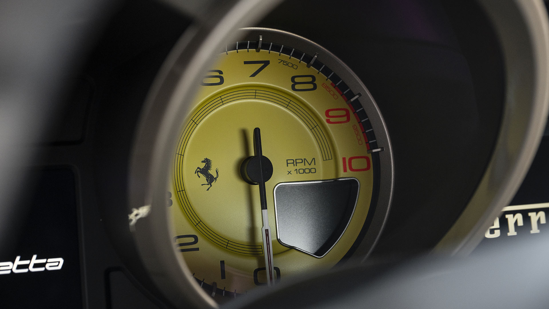 Iš arti geltono Ferrari F12berlinetta tachometro, rodančio iki 10 000 aps./min., su Ferrari logotipu centre ir raudona linija ties 8 500 aps./min., užfiksuotas prieš Broad Arrow Las Vegas 2025 renginį.