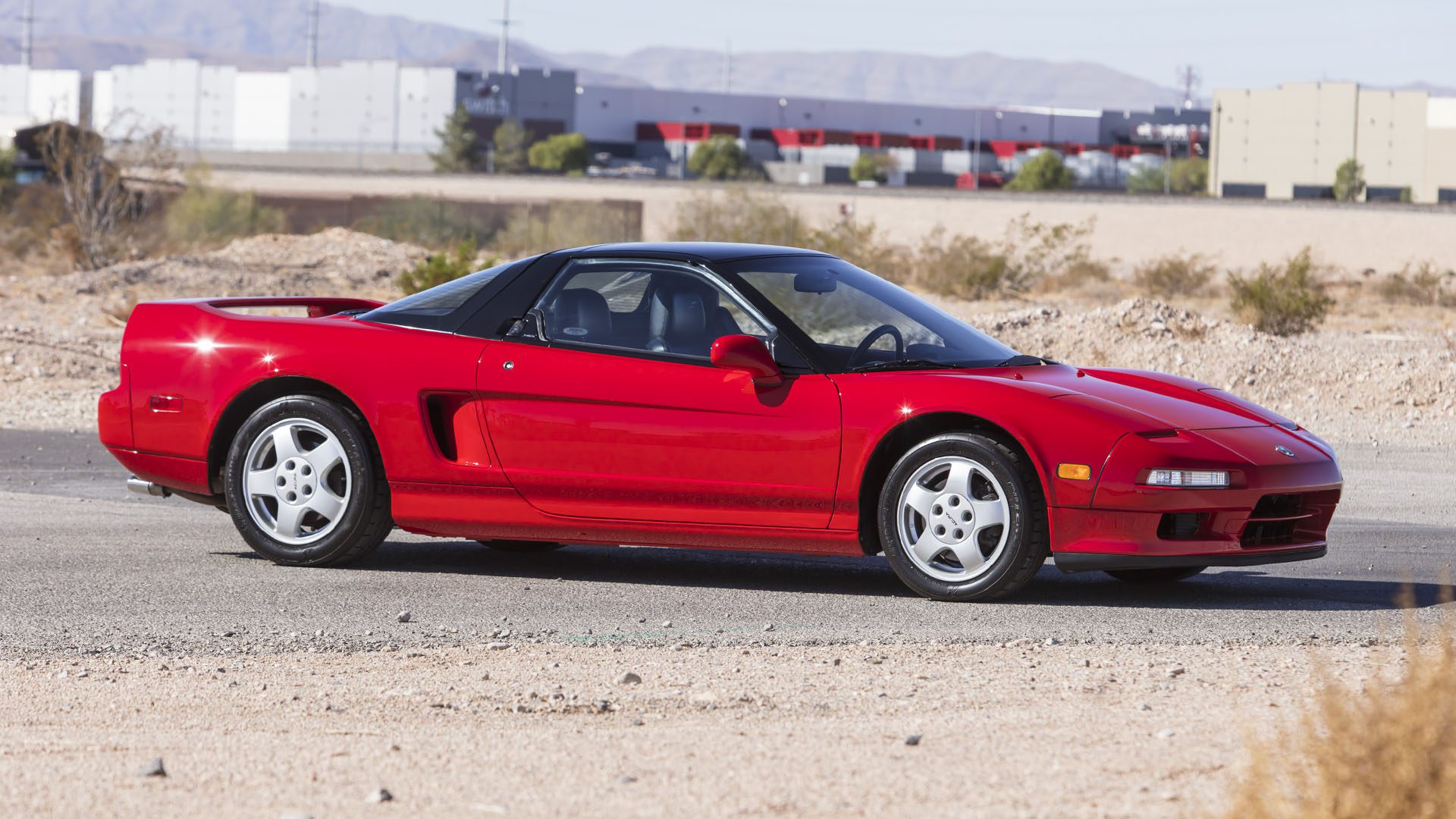 A red Ferrari SF90 Spider with a black roof is parked on a paved area near a desert, with industrial buildings and mountains in the background, making for a stunning 2025 Preview at Broad Arrow Las Vegas.